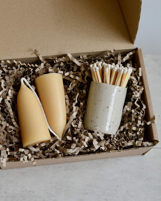 A gift box, containing a pair of beeswax candles and a ceramic match pot.