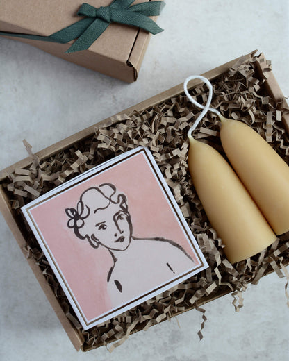 A gift box containing a pair of stubby beeswax candles and a pink matchbox with the illustration of a woman.