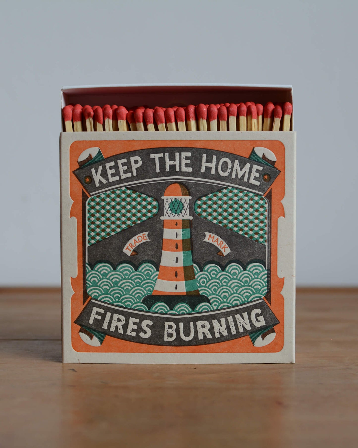 A matchbox with an orange and green illustration and lettering which reads 'Keep the home fires burning'.