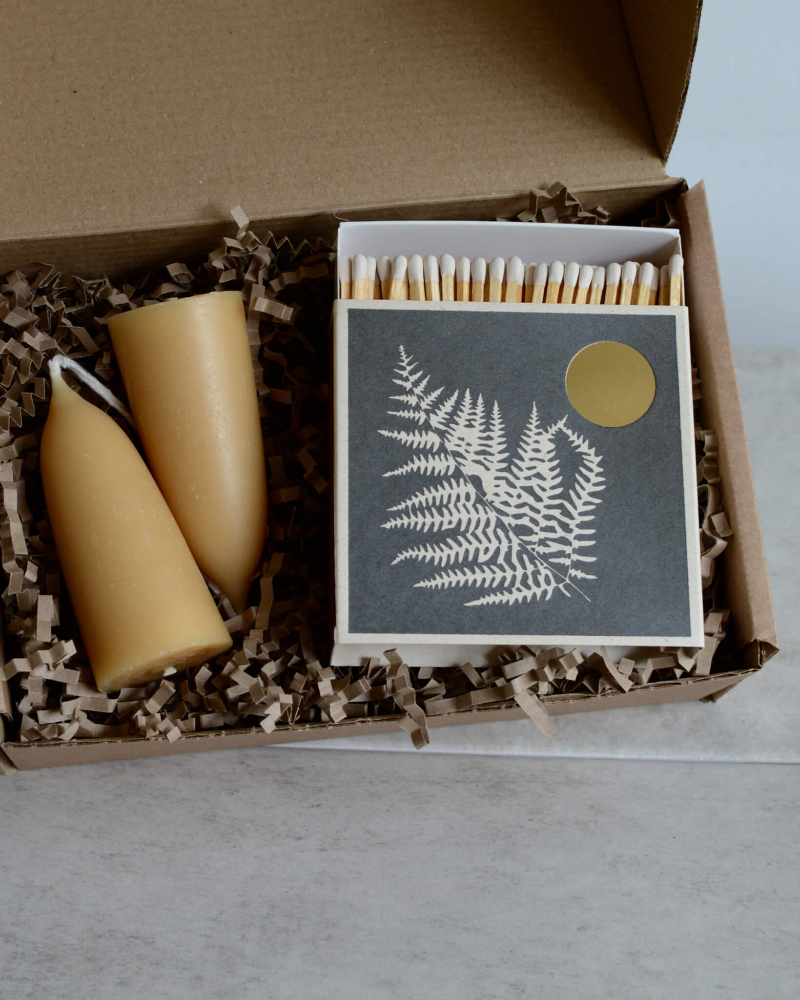 A gift box containing a pair of stubby beeswax candles and a grey matchbox with the illustration of a fern leaf.