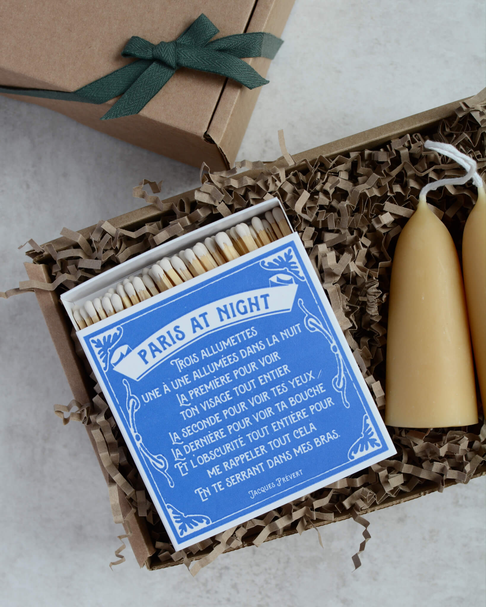 A gift box containing a pair of stubby beeswax candles and a blue matchbox with white lettering.