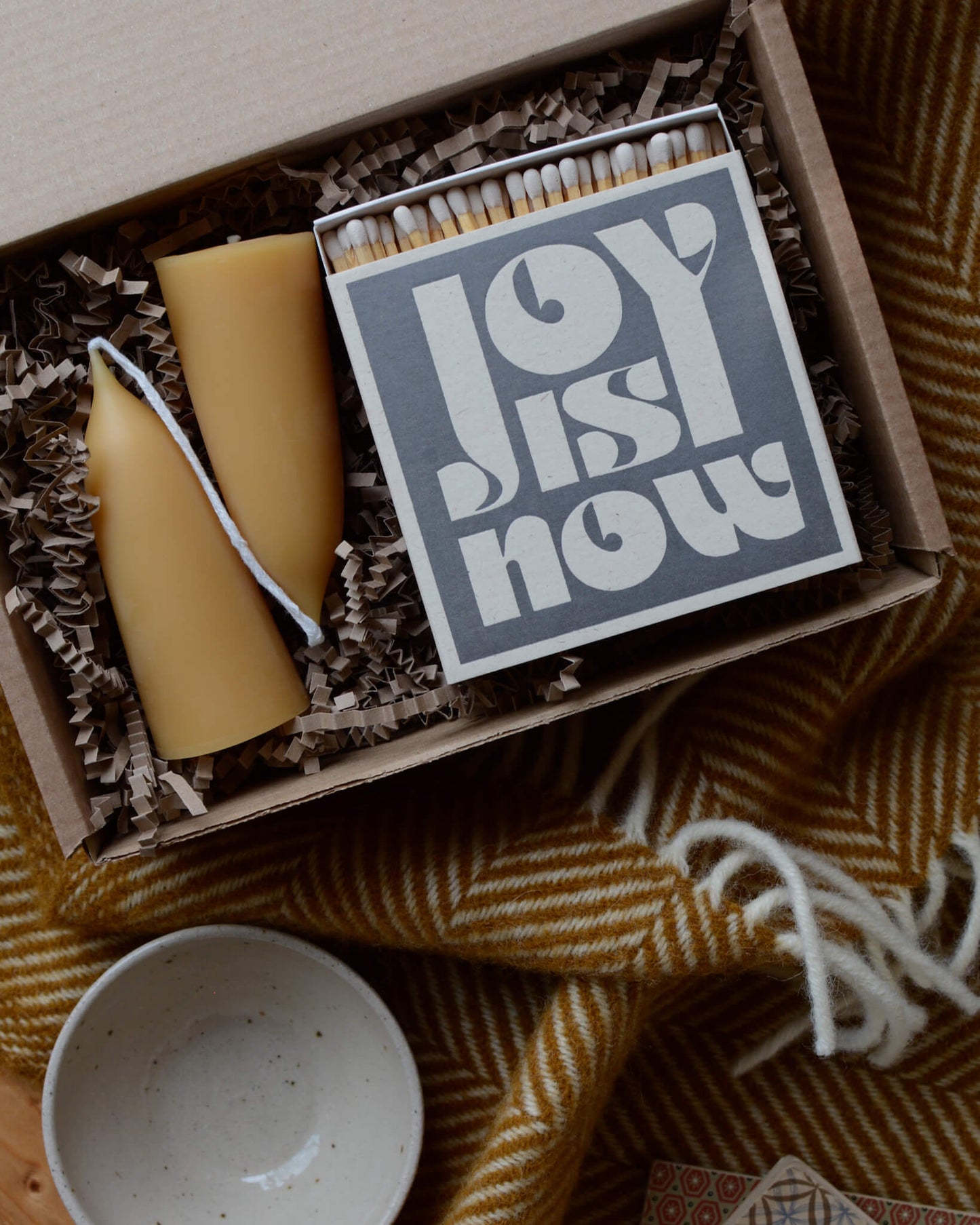 A gift box containing a pair of stubby beeswax candles and a grey matchbox with white lettering, surrounded by a mustard wool blanket.