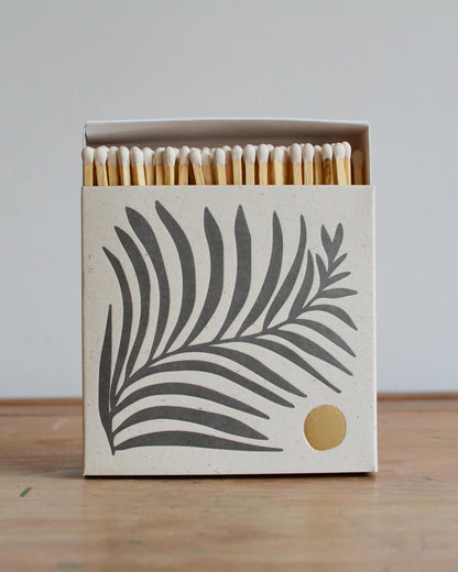 A white matchbox with a fern leaf illustration and gold leaf detailing.