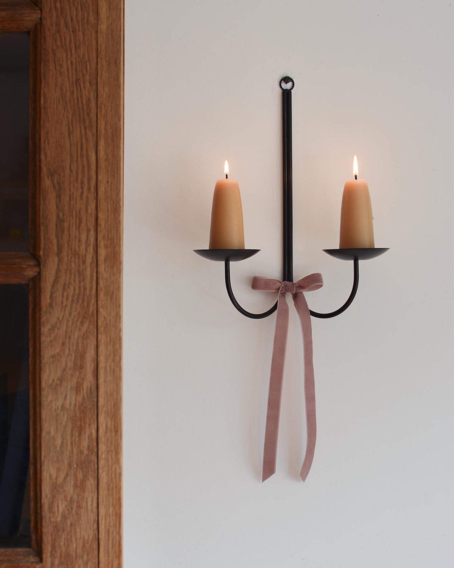 A pair of a stubby beeswax candles on a black metal wall sconce, with a pink velvet bow beneath.