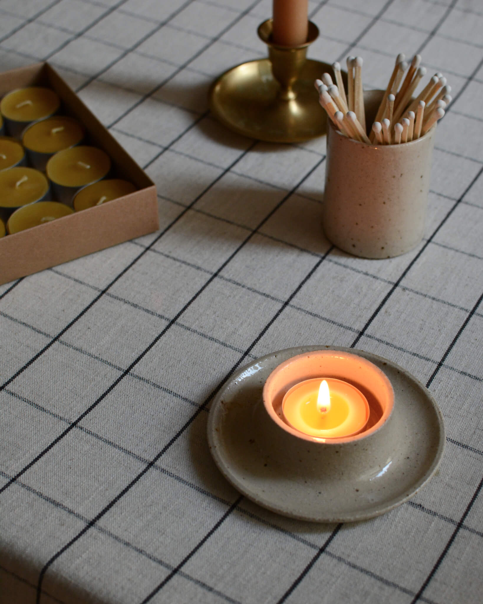 A lit beeswax tea light in a ceramic holder, on a checked tablecloth on top of a dinner table.