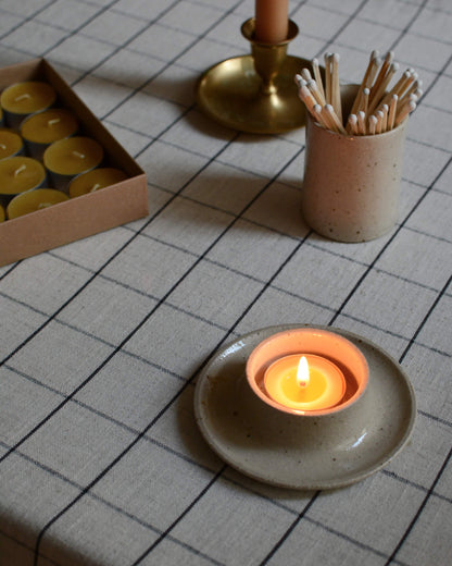 A lit beeswax tea light in a ceramic holder, on a checked tablecloth on top of a dinner table.