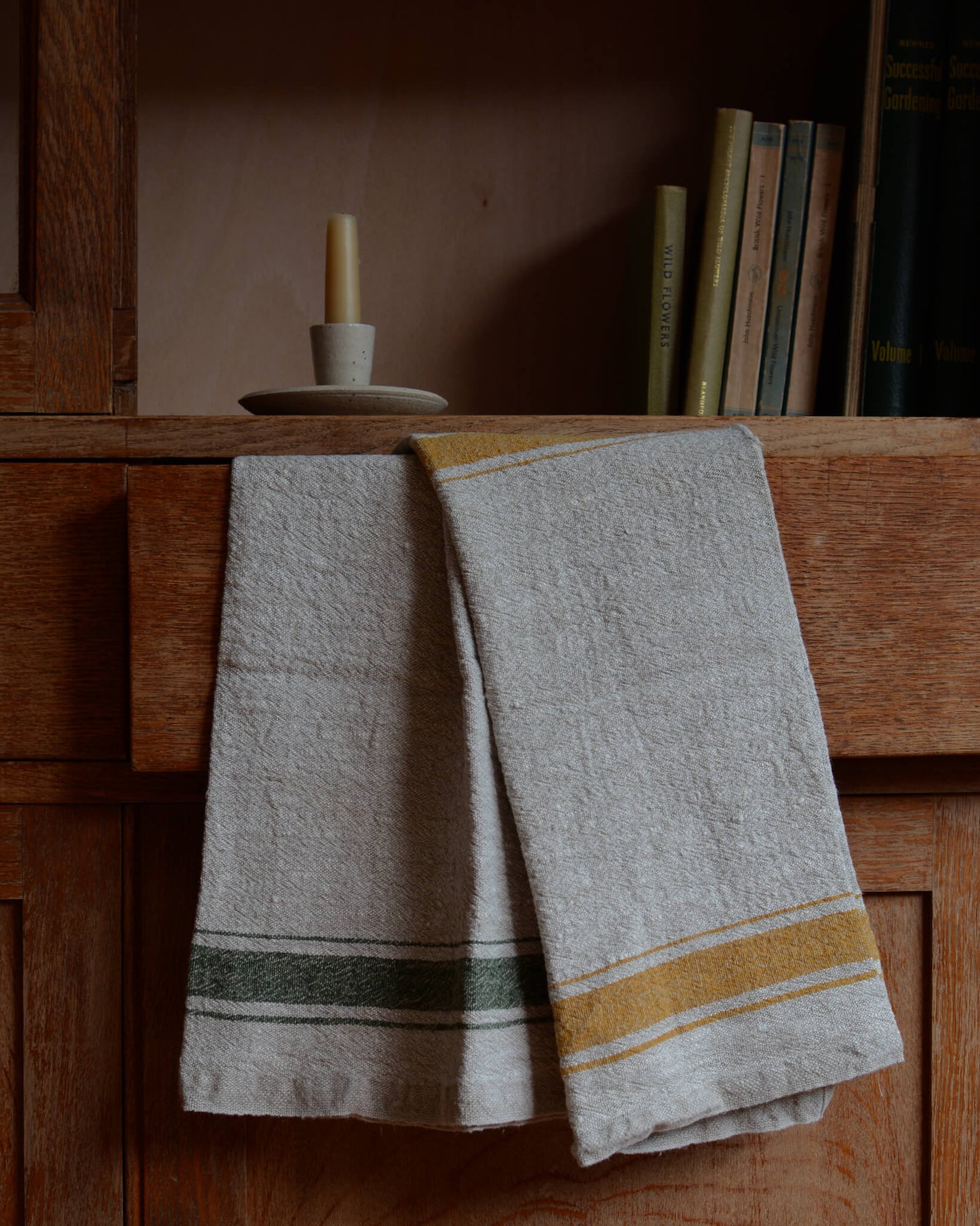 An oatmeal coloured ceramic dinner candle holder on a shelf, next to a pair of linen tea towels.