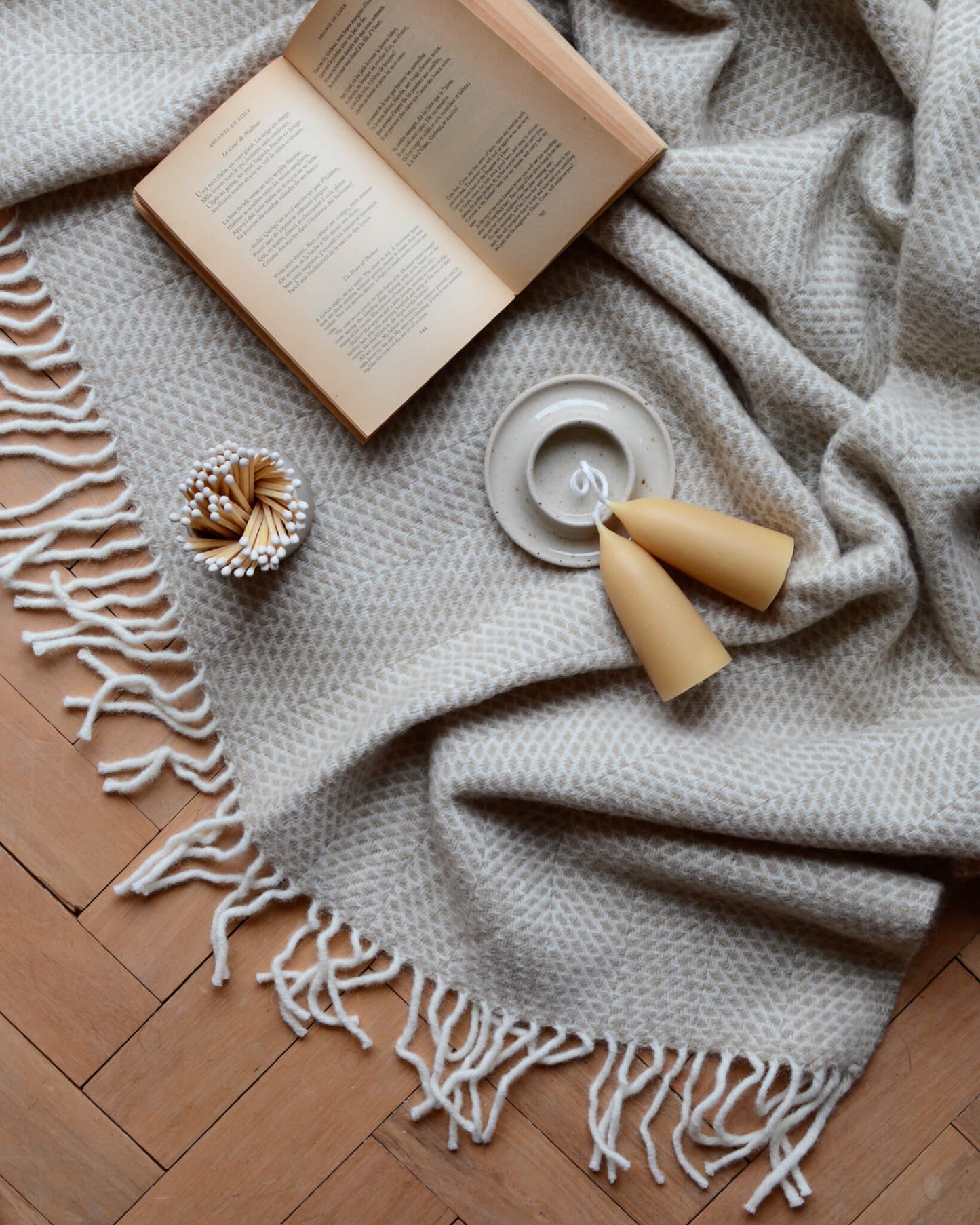 A ceramic match pot with matches, candles with a candle holder and an open book; on top of a woollen blanket.