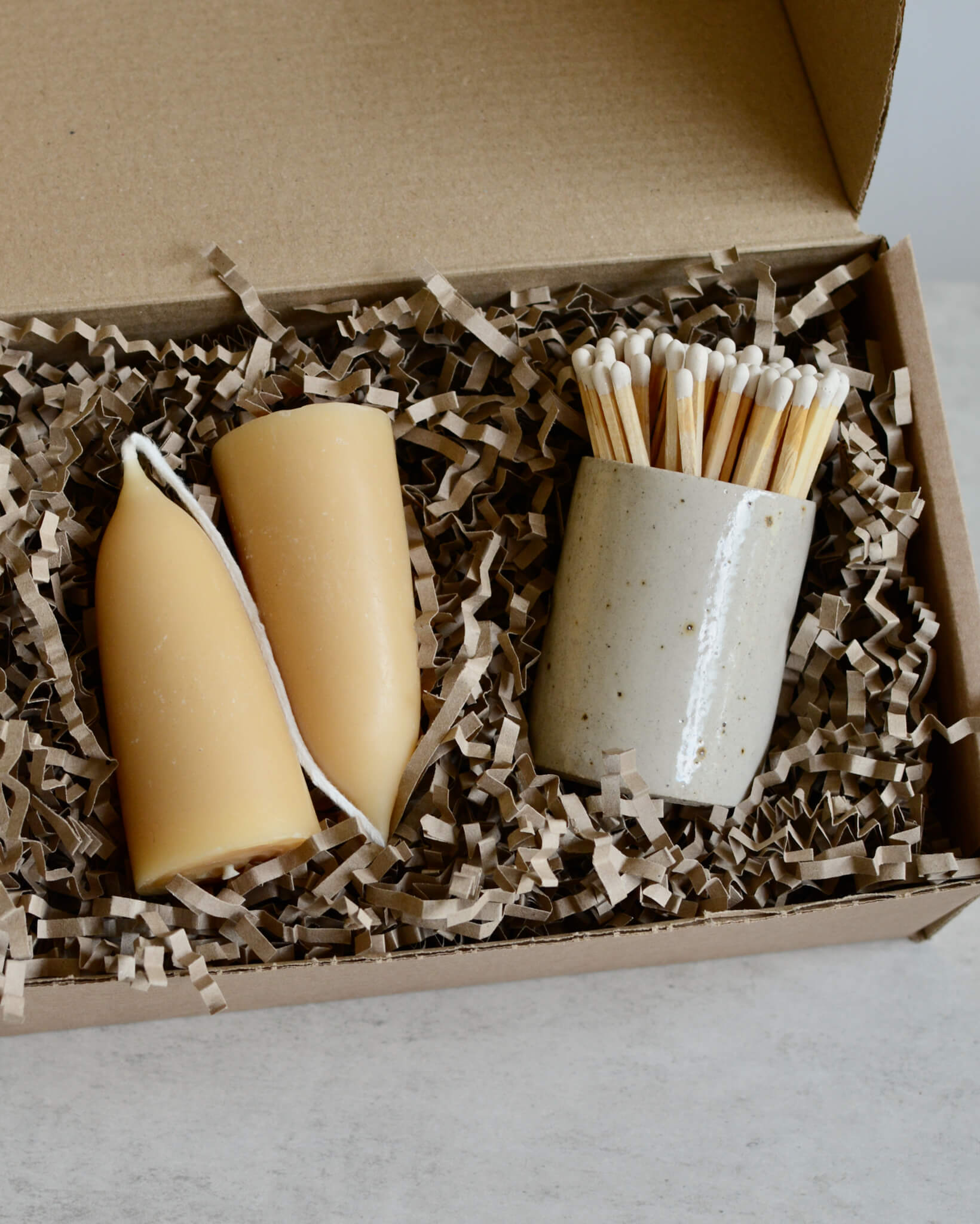 An oatmeal ceramic match pot with matches and a pair of beeswax candles, in a gift box.