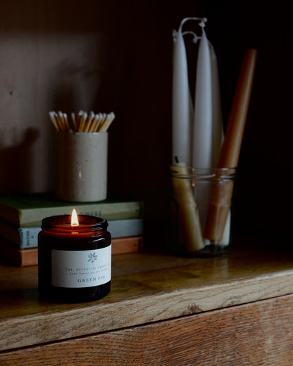 A lit candle, next to an oatmeal ceramic match pot with matches and a selection of dinner candles.