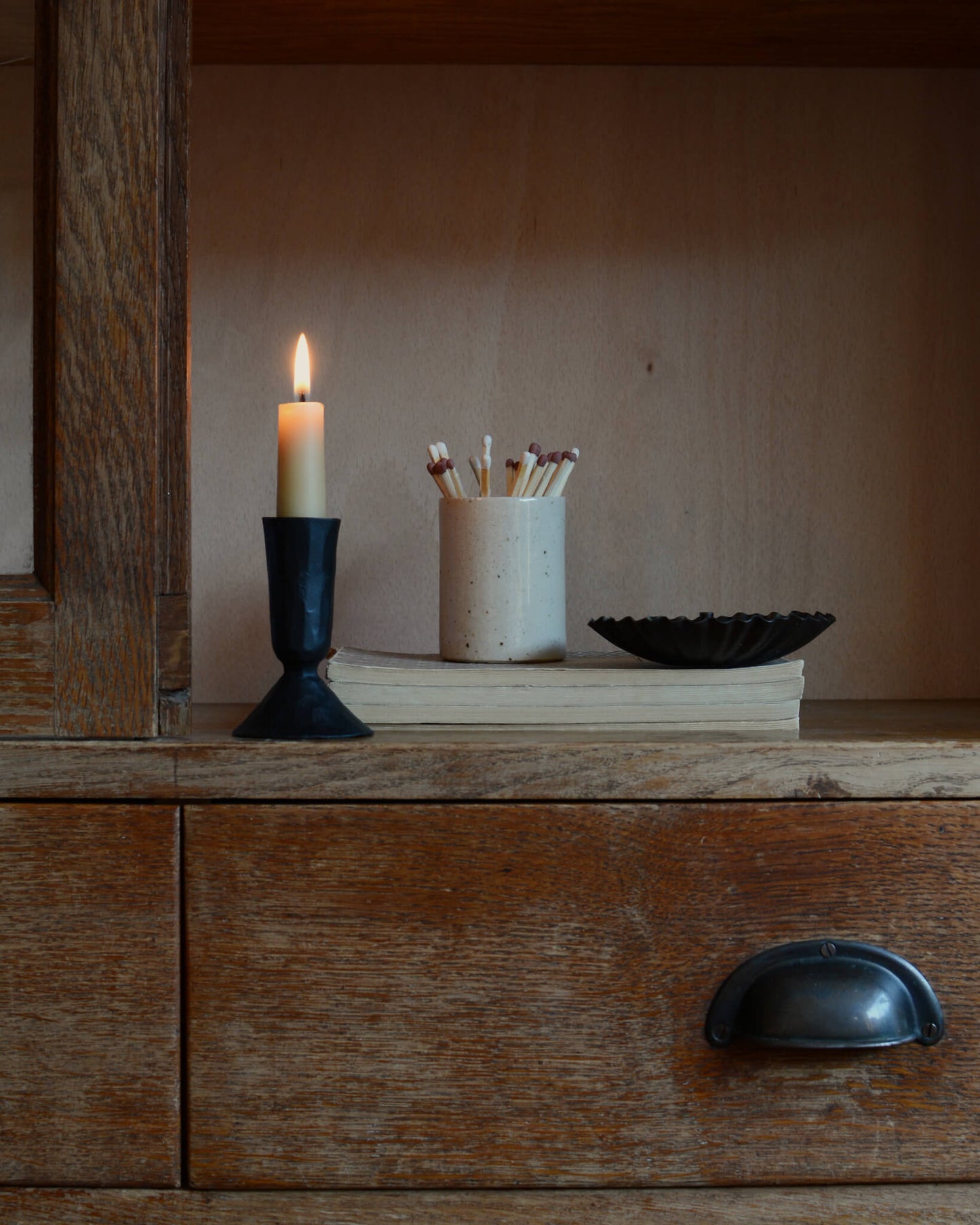 An oatmeal ceramic match pot with matches, alongside a lit beeswax candle n a black holder.