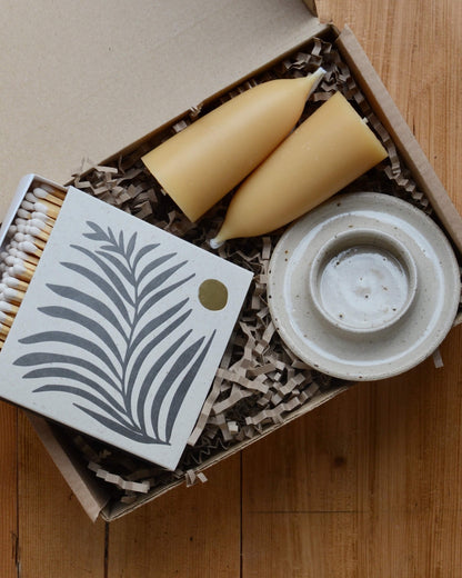 A gift box with an oatmeal ceramic candle holder and beeswax candles and a box of matches.