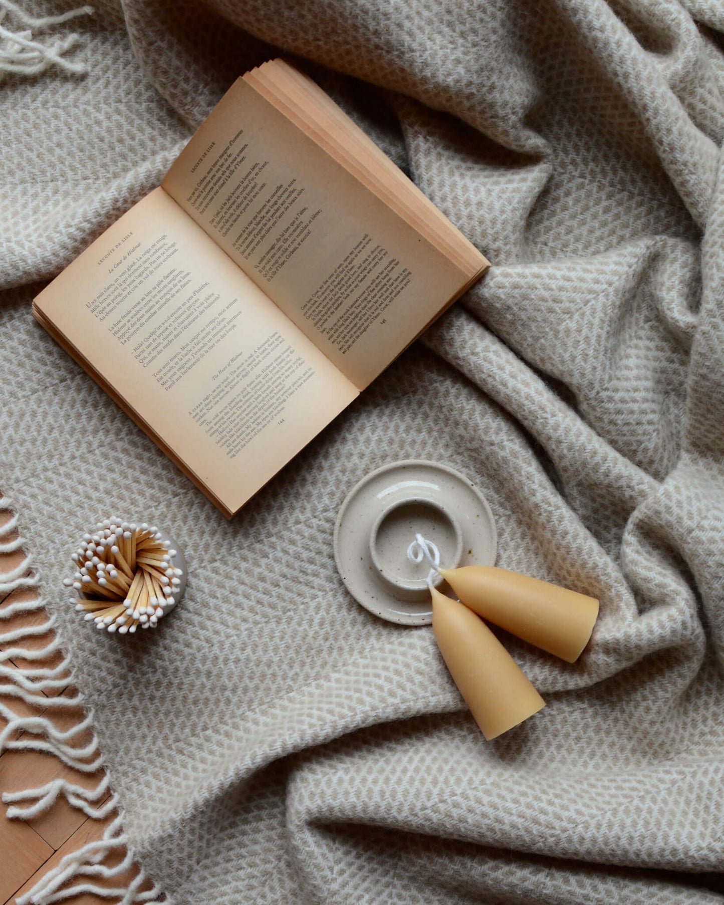 An oatmeal ceramic candle holder, beeswax candles and matches, on top of a wool blanket.