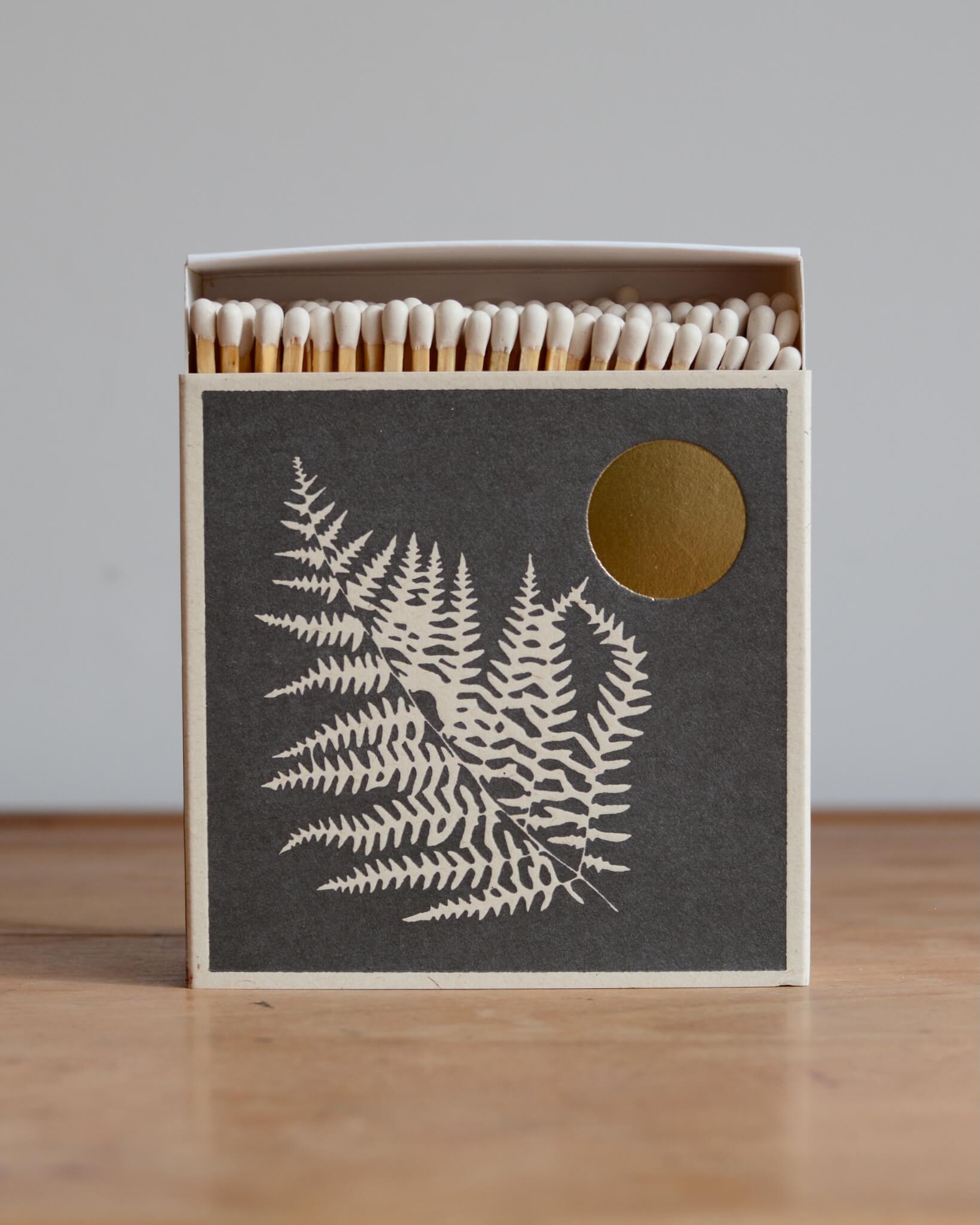 Dark grey match box with white fern leaf illustration and gold leaf moon.