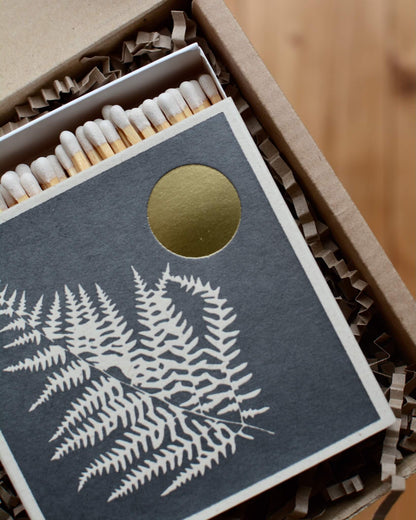 Dark grey match box with white fern leaf illustration and gold leaf moon.