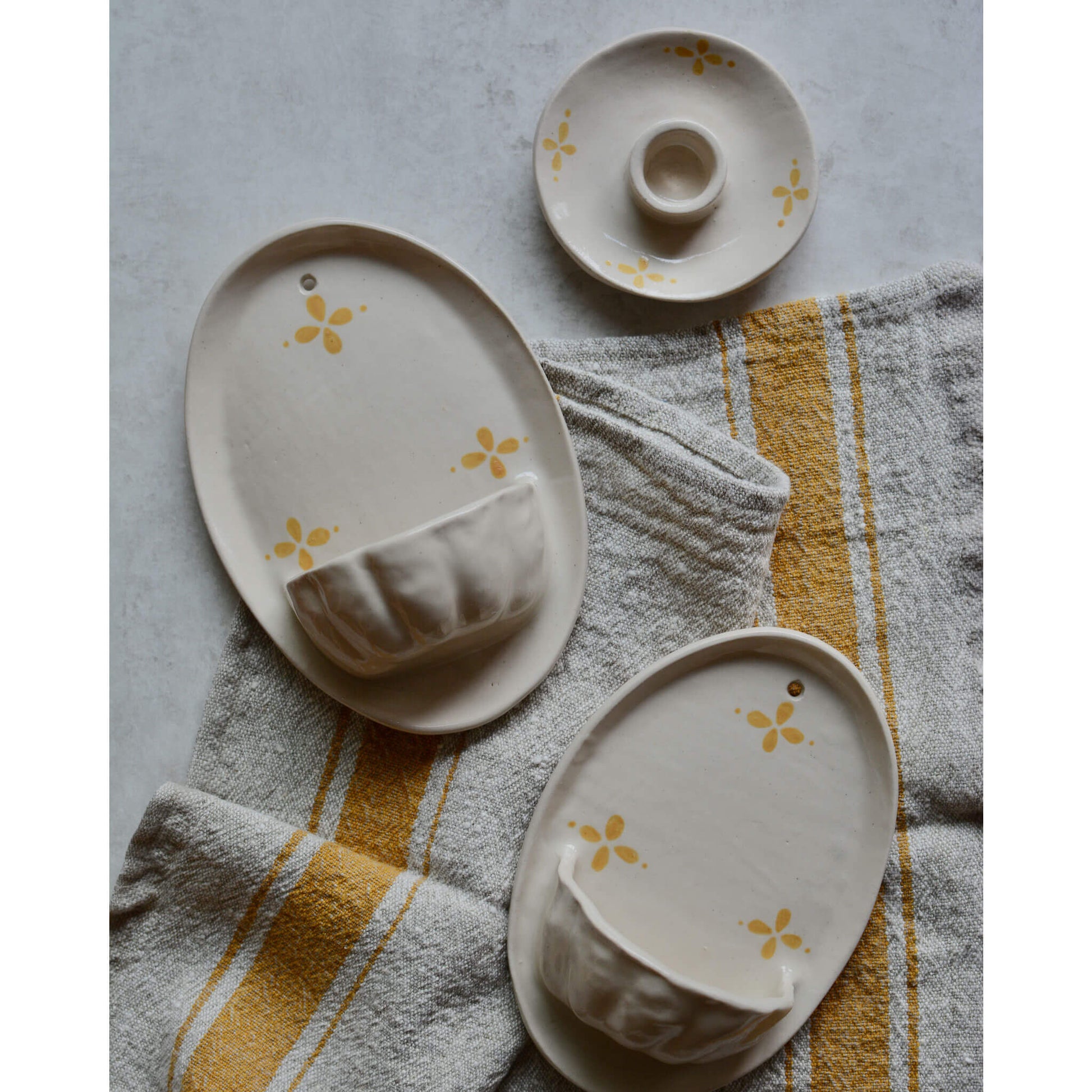 A flatlay image of cream ceramic pieces with yellow floral pattern, on top of a yellow striped tea towel.