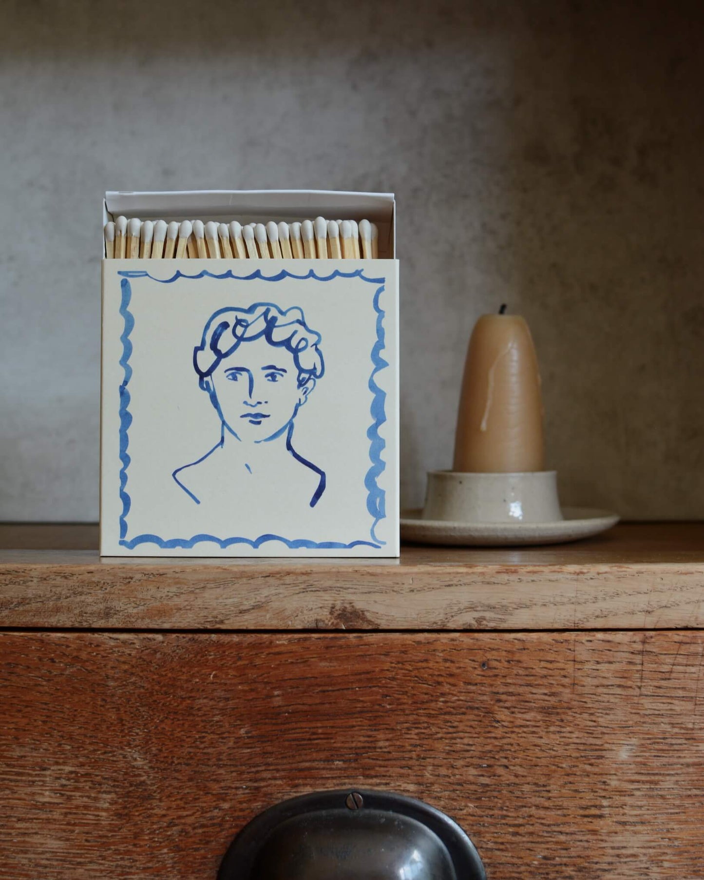 Illustrated square match box, next to a beeswax candle in a ceramic candle holder.