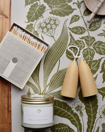 A green botanical print, depicting Spring plants, with beeswax candles and a match box.