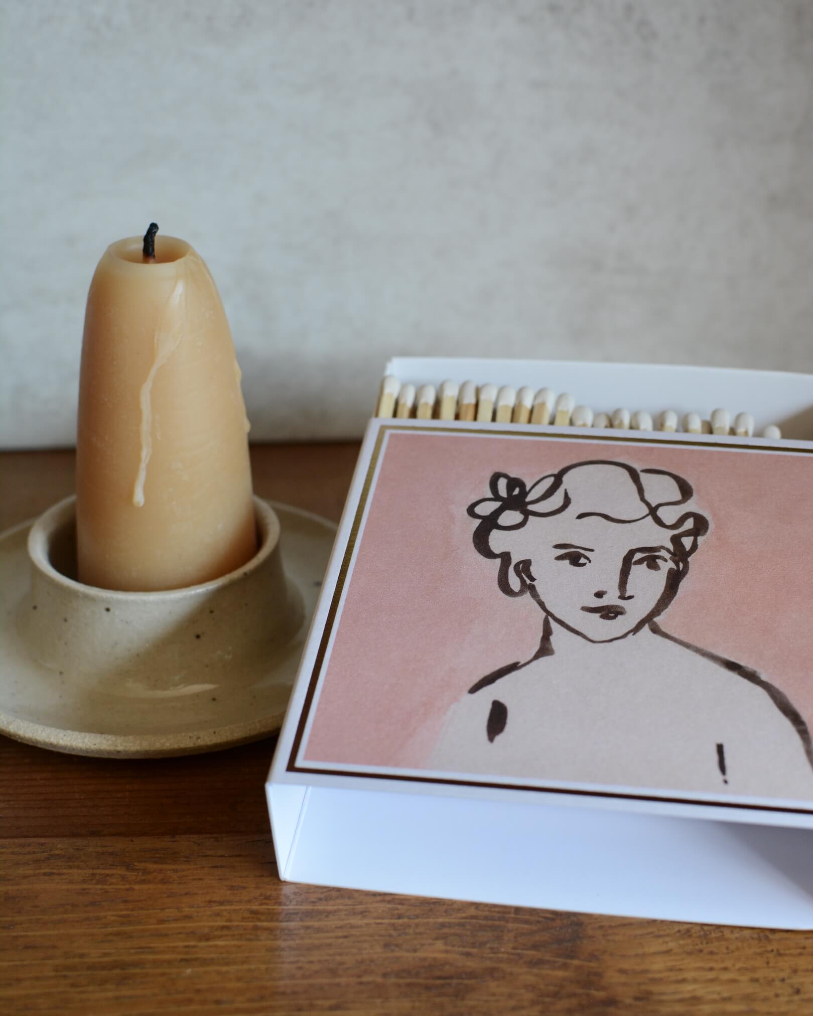 Pink matchbox featuring an illustrated portrait of a woman, next to a beeswax candle in a ceramic candle holder.