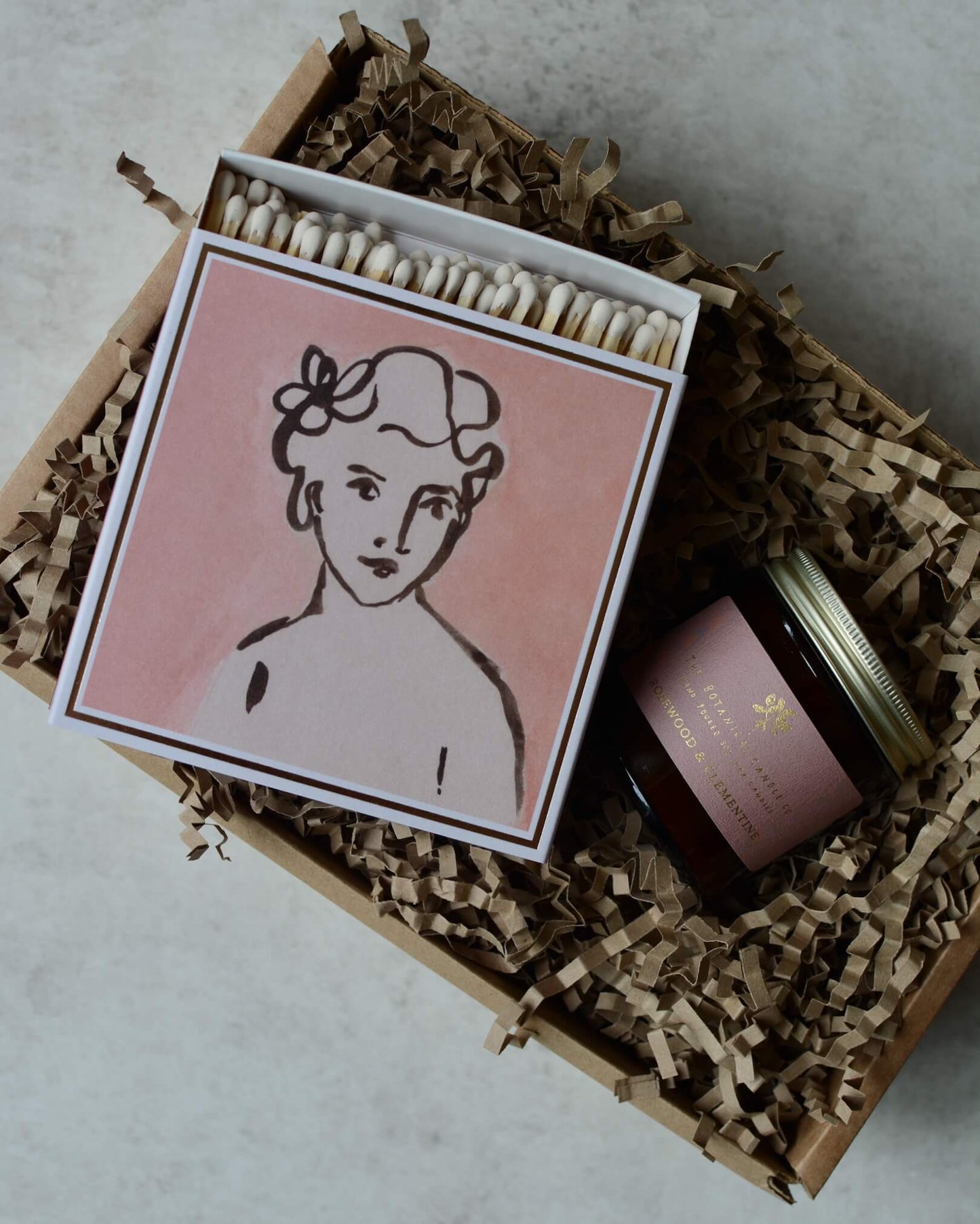 Pink matchbox featuring an illustrated portrait of a woman, next to a pink jar rose candle.