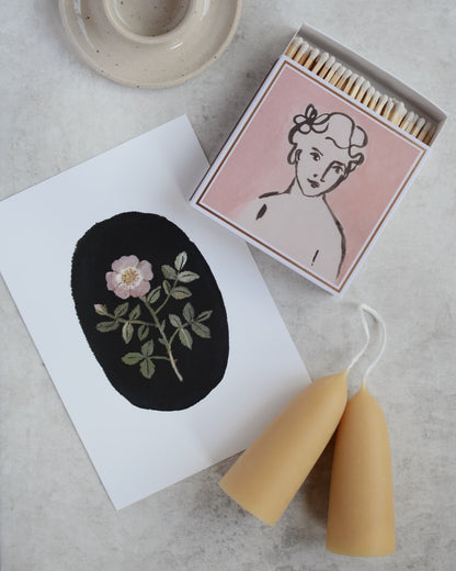 A pink matchbox featuring an illustrated portrait of a woman, a rose illustrated print and a pair of beeswax stubby candles.