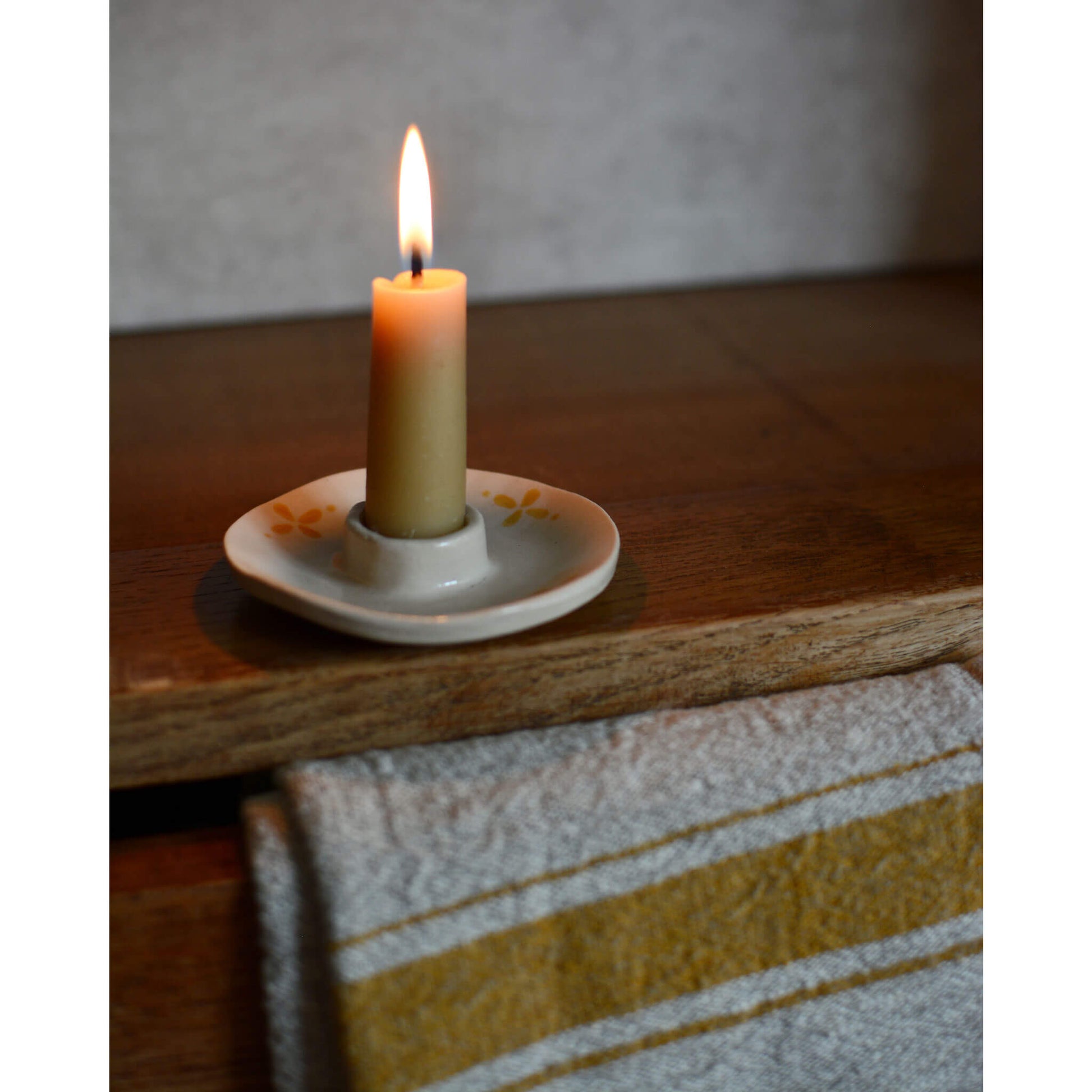 A styled image of a ceramic candle holder with a beeswax candle with flame on a wooden cabinet.