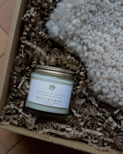 Redamancy scented candle by The Botanical Candle Co. in a gift box next to a sheepskin.