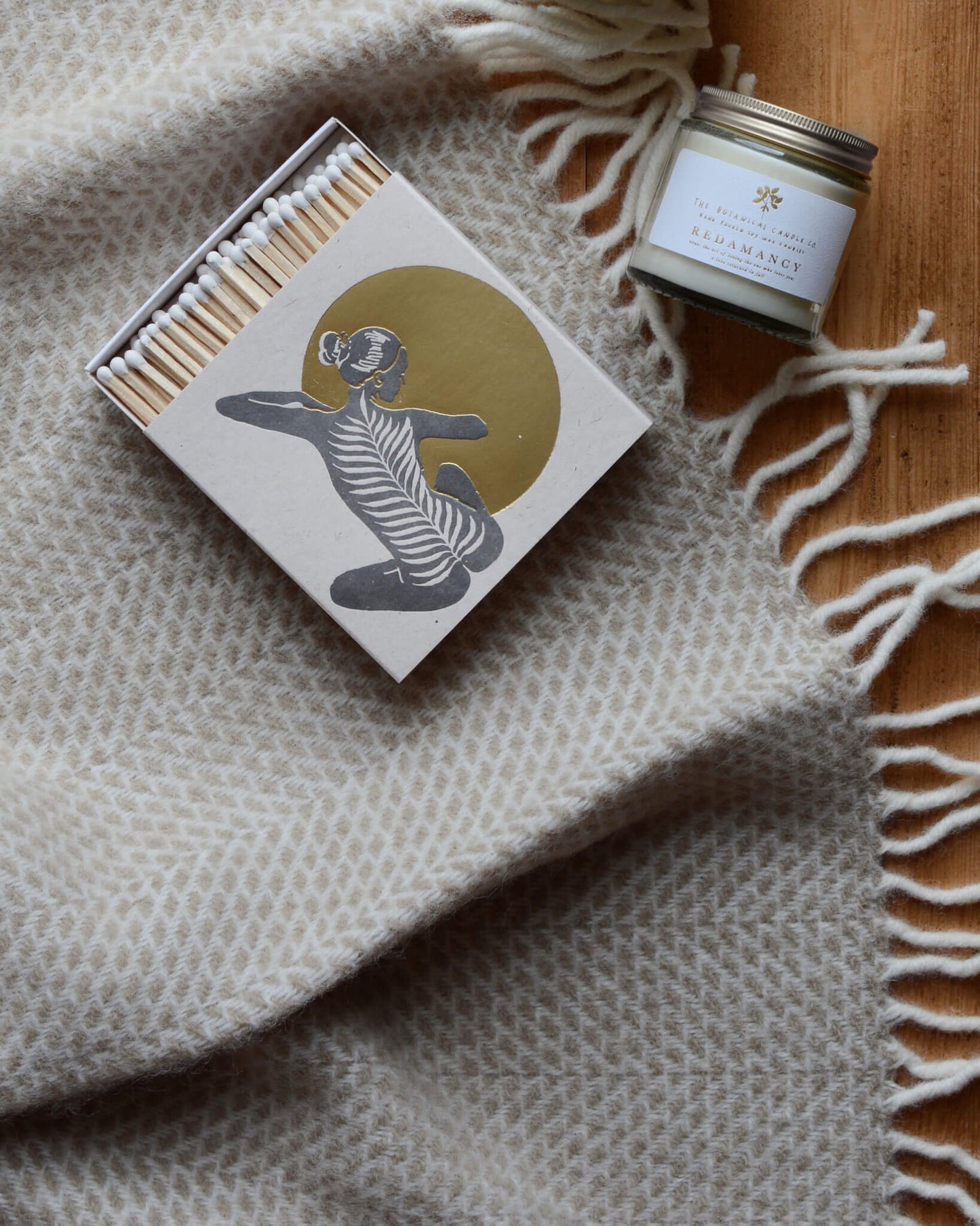 Redamancy scented candle by The Botanical Candle Co. on a woollen blanket, next to a box of matches.