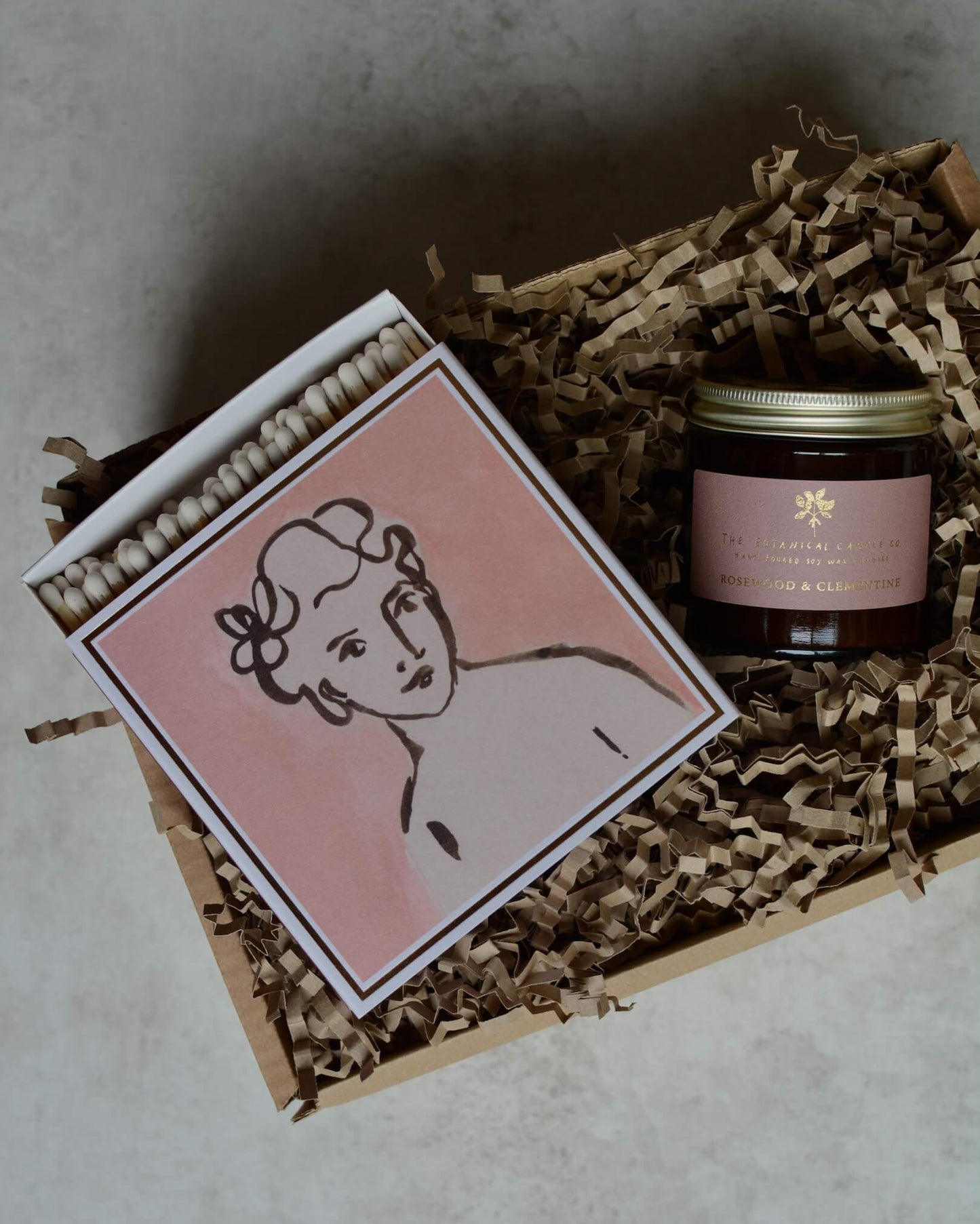 A gift box containing a pink rose candle and a pink and white illustrated box of matches.