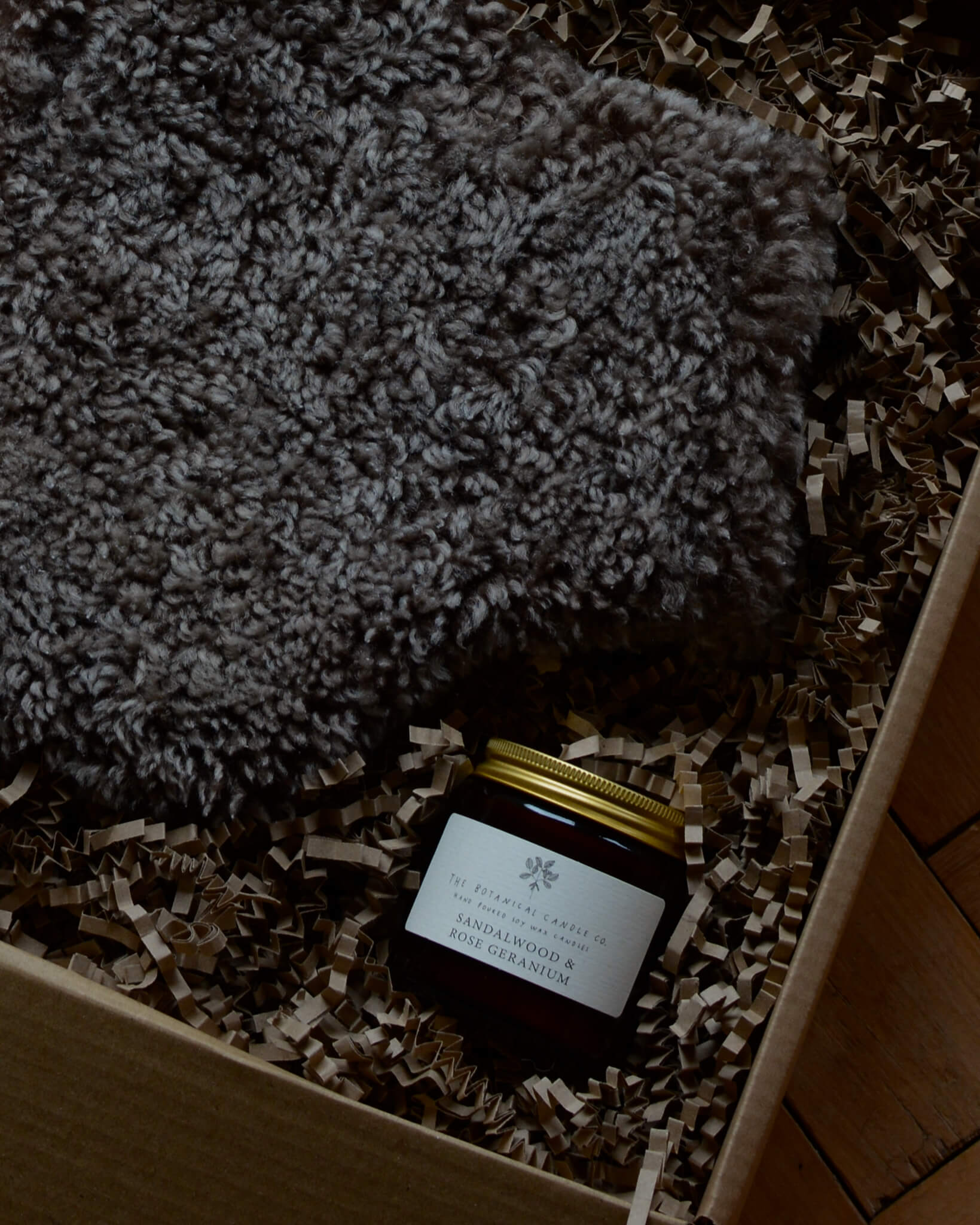 A brown sheepskin hot water bottle in a gift box, alongside a candle.