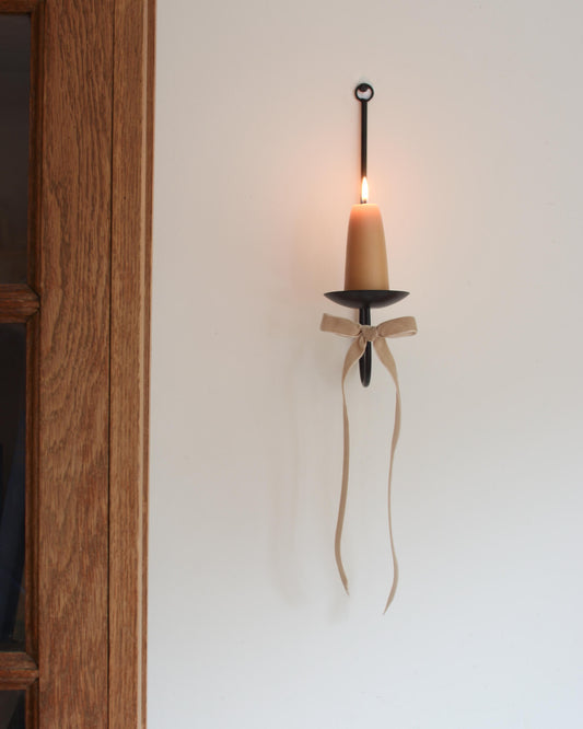 A single black metal wall sconce candle holder with a lit beeswax stubby candle and a velvet bow beneath.