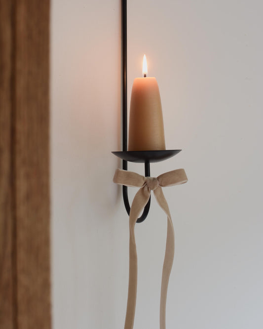 A black wall mounted candle sconce, with a beeswax candle and a beige velvet bow. 