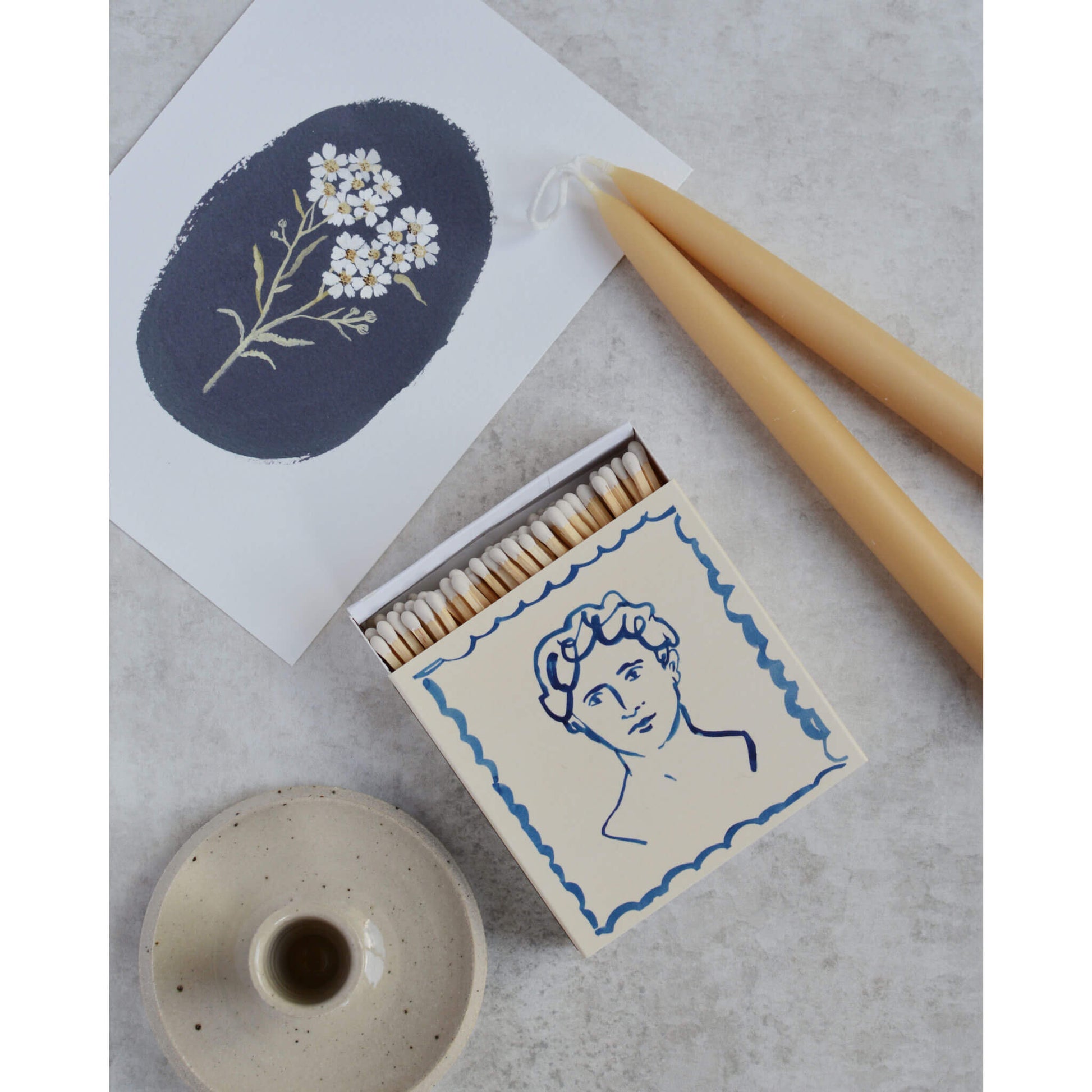 Illustrated square match box, next to a botanical illustration and beeswax candles.