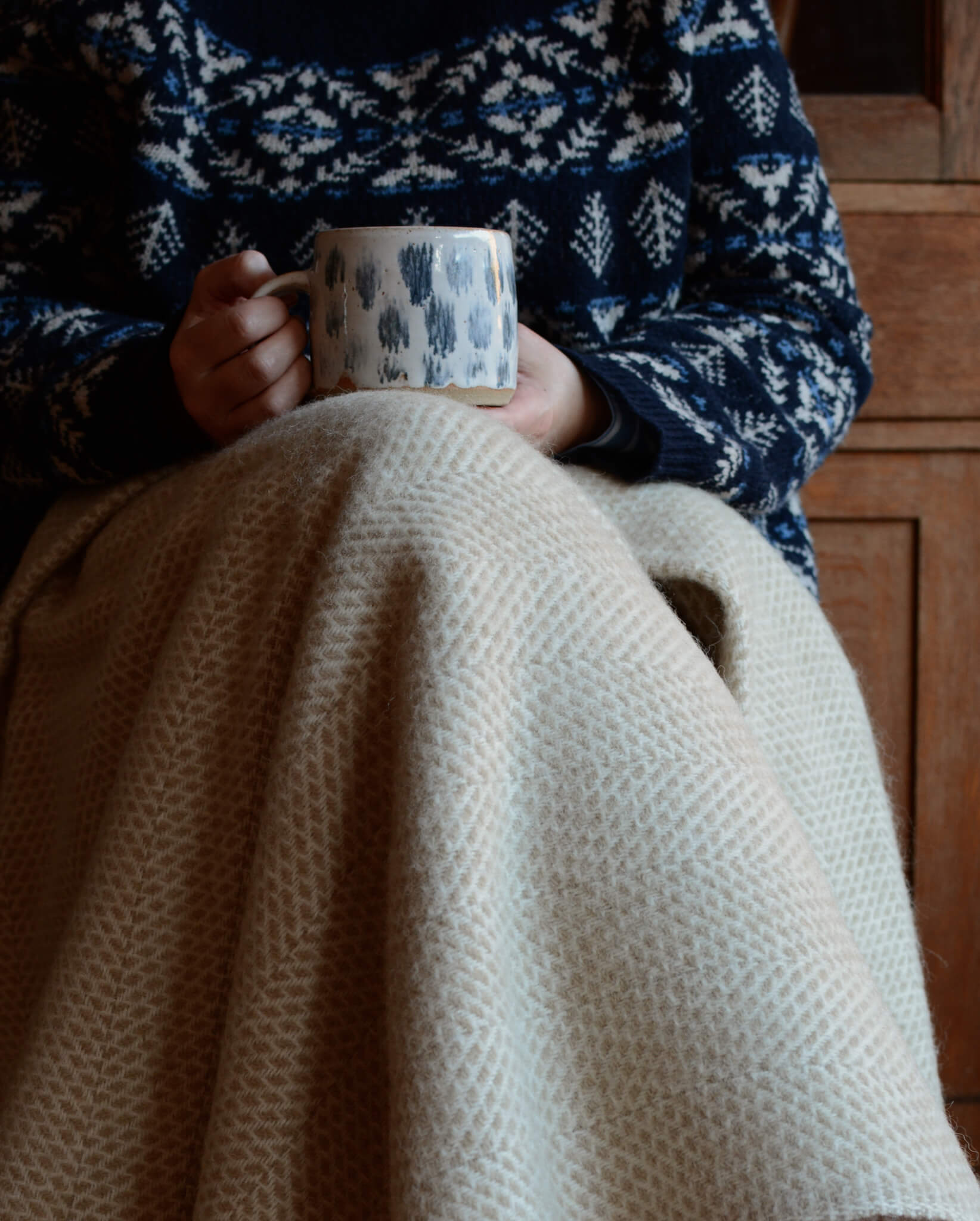 An oatmeal beehive pattern blanket, on the lap of a woman, holding a coffee.