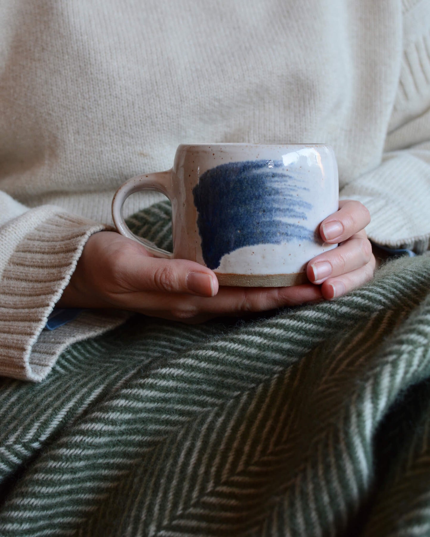 A woman holding a coffee cup with a green woollen herringbone blanket on her lap.