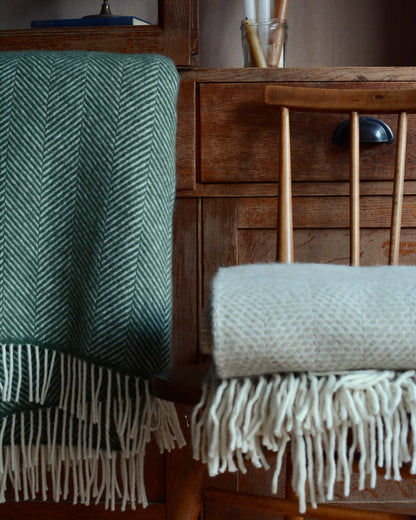 A green woollen herringbone blanket next to an oatmeal blanket, in front of a wooden cabinet. 