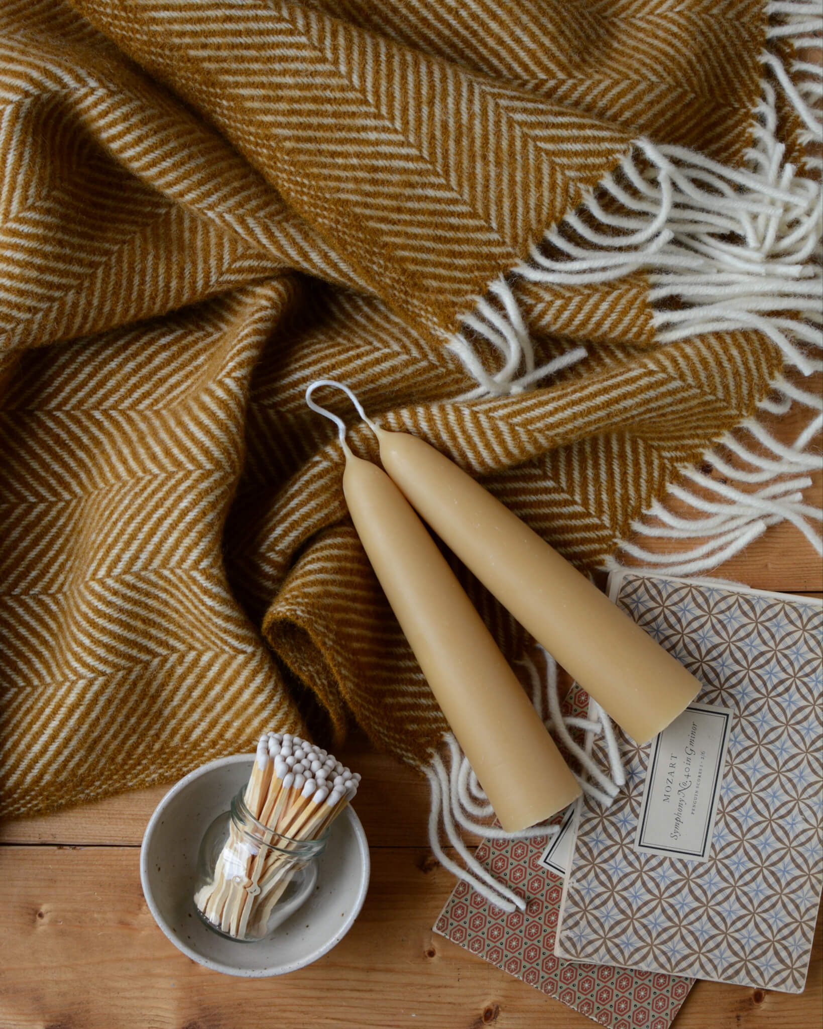 A mustard yellow wool herringbone blanket with beeswax candles and patterned books.