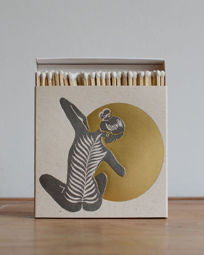 A white match box featuring a yoga figure in front of a shiny gold moon. 